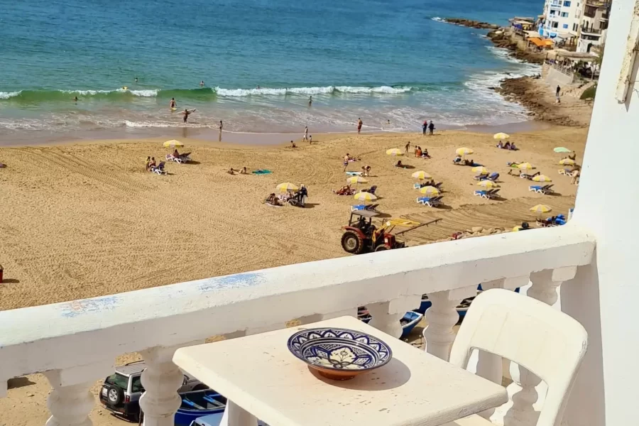Beachfront apartment terrace overlooking Taghazout beach with surfers and swimmers in the water below.