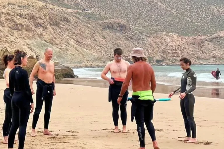 Surf instructor briefing a group of students on the sandy beach of Imsouane with dramatic cliff faces in the background.