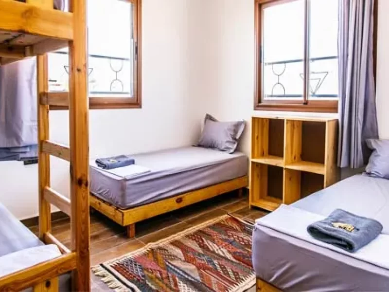 Shared bedroom with wooden single bed, storage shelving, and windows overlooking Taghazout village.