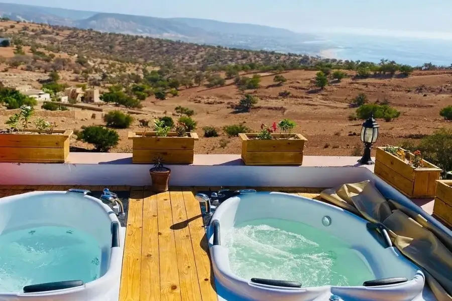 Terrasse mit zwei Whirlpools, Holzboden und bepflanzten Kästen mit Blick auf die Küstenhügel und das Meer von Taghazout.