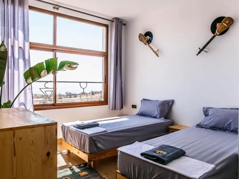 Twin beds with blue bedding in a bright room at Sun House Morocco with windows overlooking Taghazout landscape.