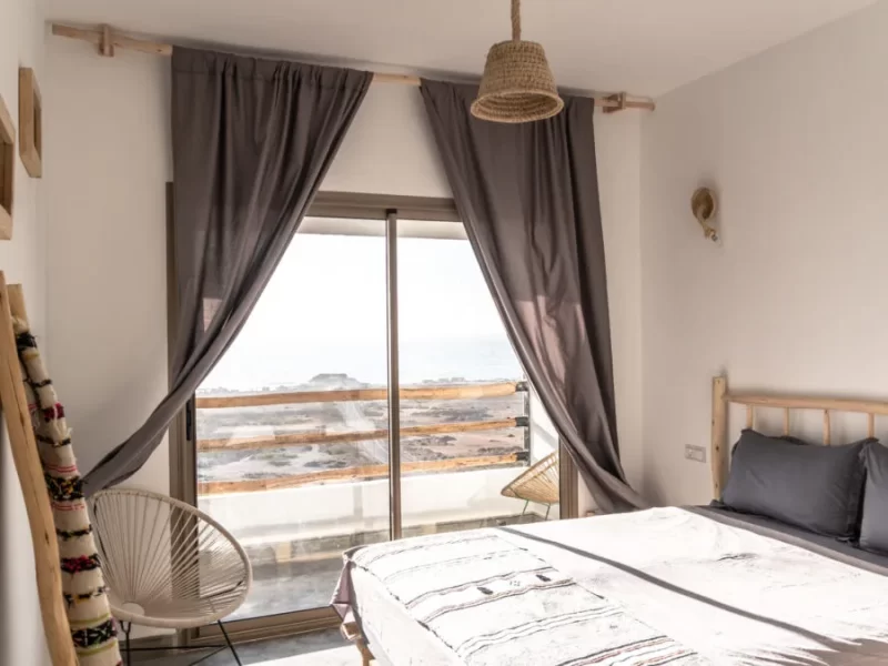 Double bedroom with direct ocean view through glass terrace door, featuring white bedding, dark curtains, and woven pendant lamp.