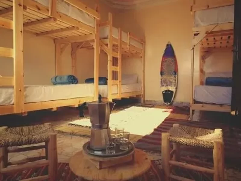 Chambre dortoir partagée avec lits superposés en bois, tapis tissés au sol et présentation de planche de surf colorée à Imsouane Soul Lodge.