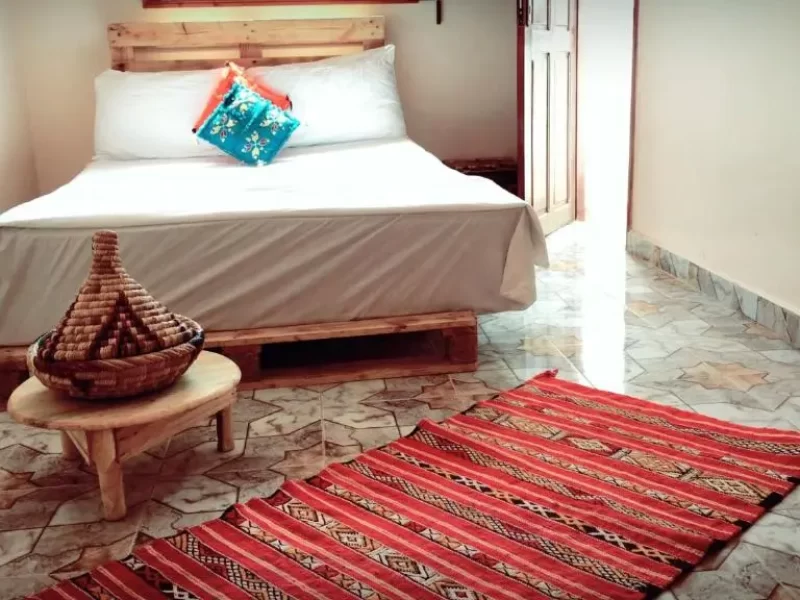 Chambre double à Imsouane Soul Lodge avec lit en bois à linge blanc, coussins bleus et corail, et tapis traditionnel rouge à rayures sur sol carrelé.