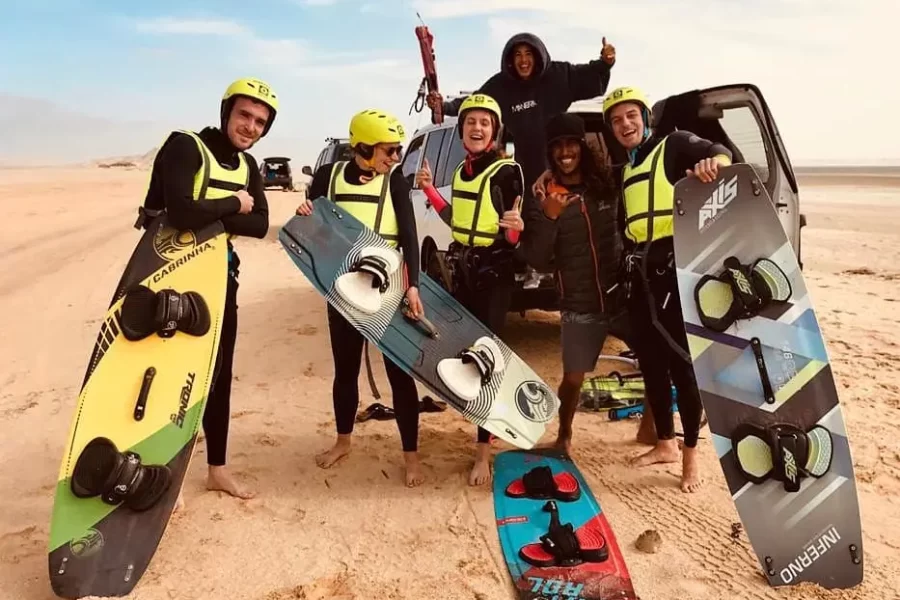 Group of six people in wetsuits and safety helmets display kiteboarding equipment on a sandy beach in Dakhla.
