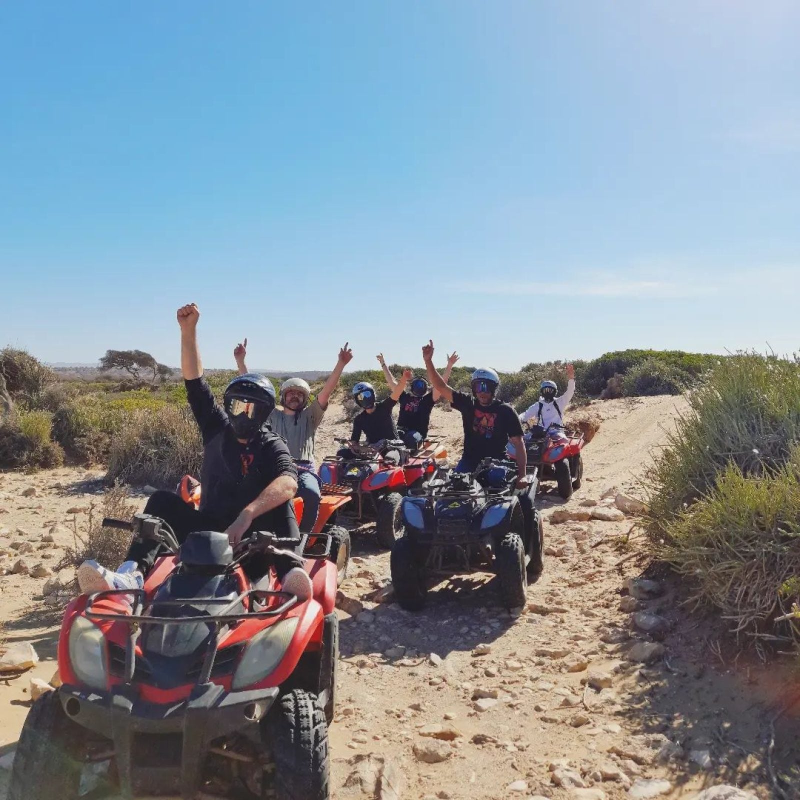 Eine Gruppe von Personen auf roten und blauen Quads feiert mit erhobenen Fäusten in einer sandigen Wüstenlandschaft mit spärlicher Vegetation.