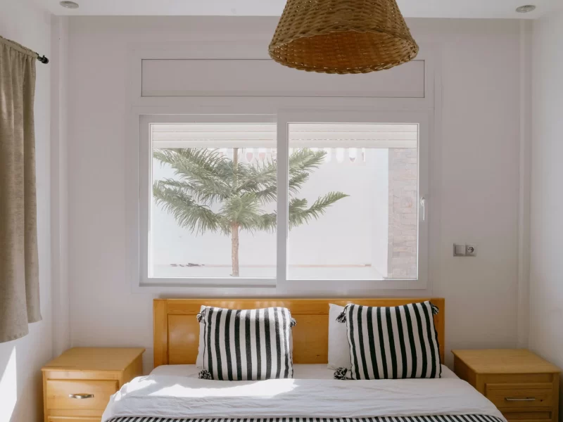 Double room at Surf Villa Morocco with white bedding, wooden headboard, striped cushions, and windows overlooking palm trees and ocean.