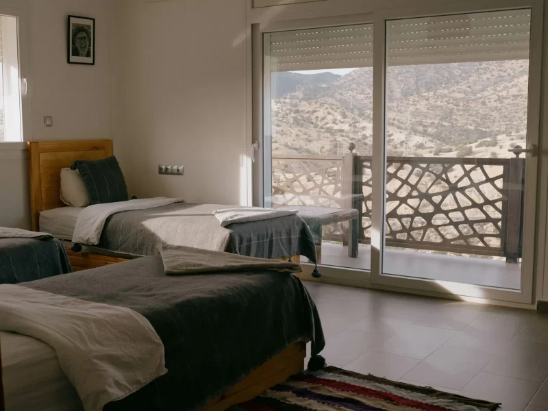 Triple shared room with wooden-framed beds, striped rug, and panoramic terrace overlooking arid mountains.