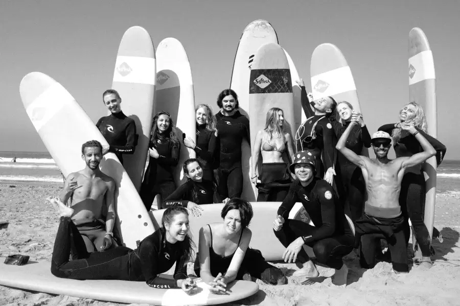 Surfing group of mixed ages and genders posed with multiple white surfboards on sand at Taghazout beach.