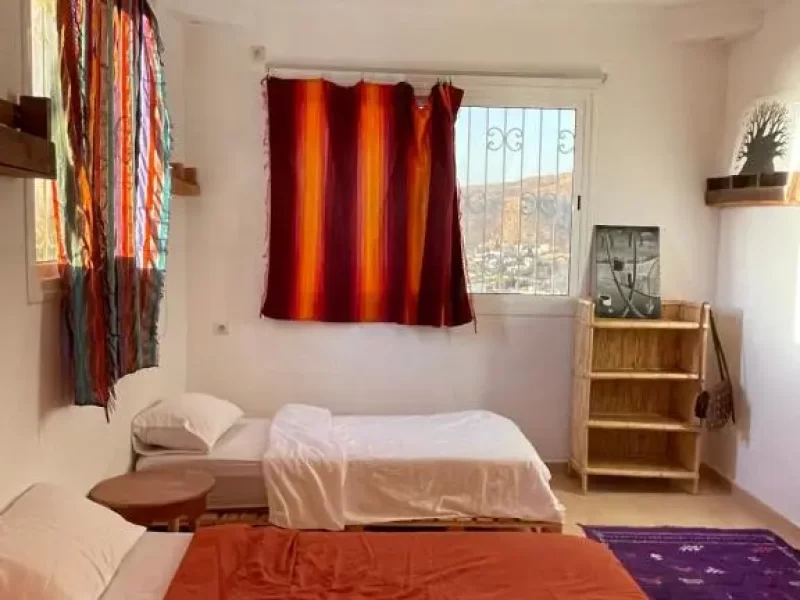 Triple shared bedroom at Kosa Surf Camp featuring two beds, wooden storage shelves, woven pendant light, and curtained window overlooking Taghazout valley.