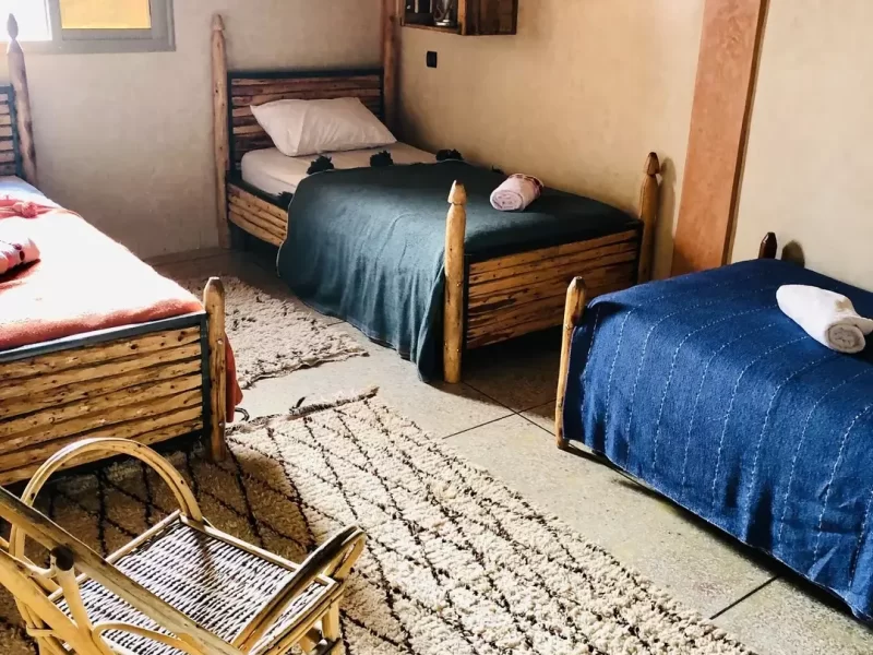 Chambre triple partagée à Tamazirt by Amayour avec trois lits simples en bois, couvertures colorées et tapis tissés au sol.