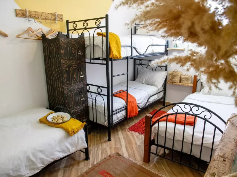 Viererzimmer-Gemeinschaftsraum im Amayour Surf Camp mit schwarzen Metall-Etagenbetten, weißer Bettwäsche und bunten Decken in Orange- und Gelbtönen.