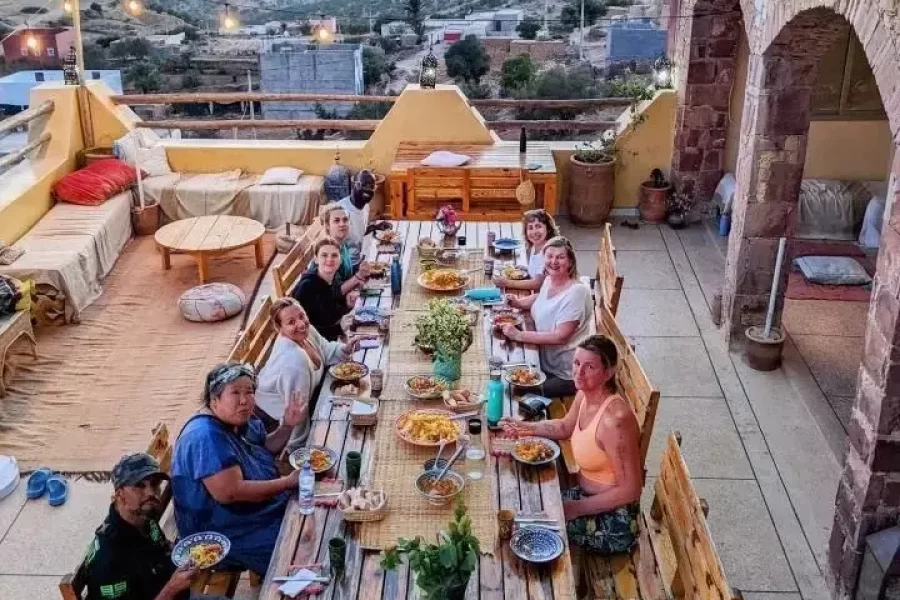 Guests gathered around a long wooden table dining on the rooftop terrace of Tamazirt by Amayour, with the Taghazout valley and hillside town visible at dusk.