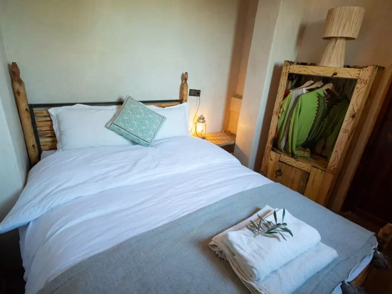Lit double avec linge blanc et oreiller turquoise dans une chambre minimaliste avec armoire en bois et éclairage d'ambiance.