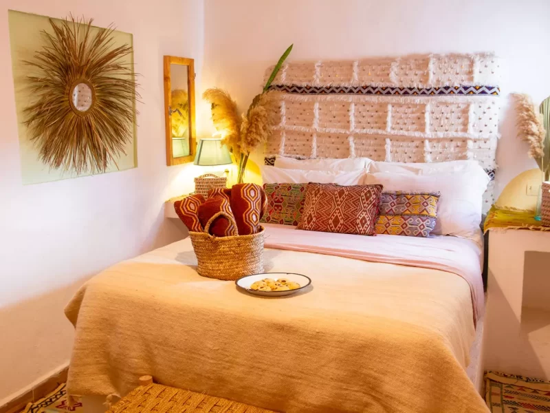 Doppelzimmer im Amayour Surf Camp mit Bett mit dekorativen gemusterten Kissen, geflochtenem Korb und warmer Akzentbeleuchtung.