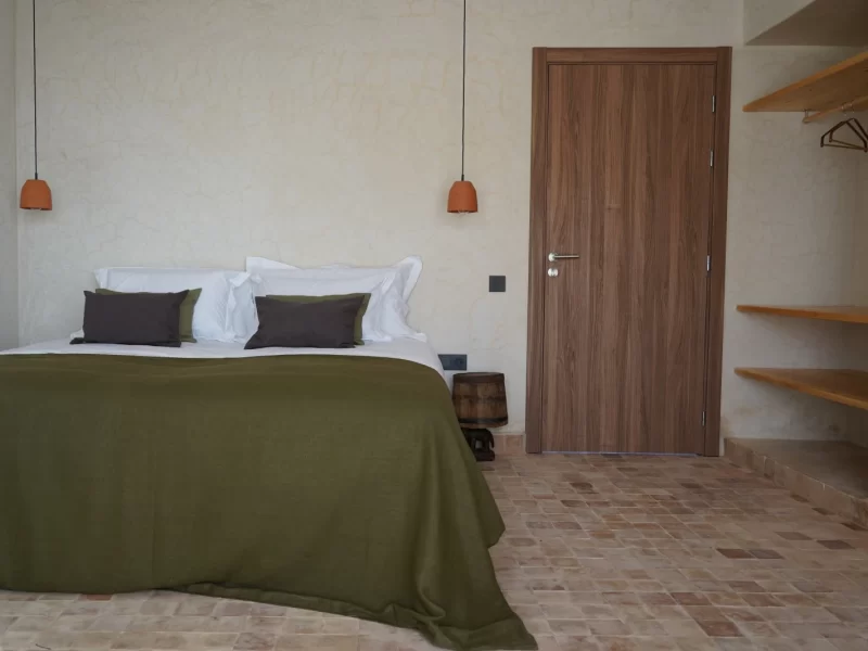 Doppelzimmer im L'Oued the Lodge mit Bett mit olivgrüner Bettwäsche, weißen Kissen, dunklen Akzenten und Holztüreingang.