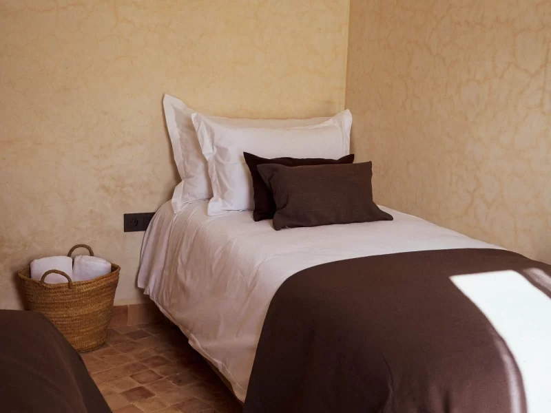 Twin-Zimmer im L'Oued the Lodge in Imsouane mit Bett mit weißen Laken, dunkelbraunen Kissen und Überwurf sowie geflochtenem Korb mit Toilettenartikeln auf Terrakotta-Fliesenboden.