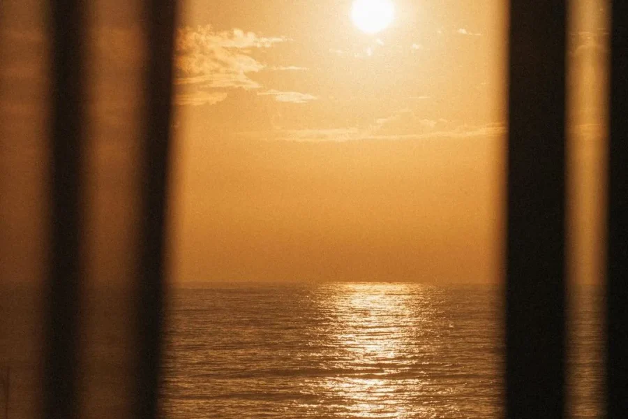 Goldener Sonnenuntergang mit heller Sonne, die sich auf ruhigem Meerwasser spiegelt, eingerahmt durch transparente Vorhänge in einem Innenraum in Imsouane.