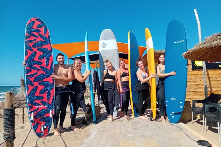 Group of eight surf students holding various colored boards on sand at Jack Beach, Casablanca with shack and orange canopy in background.