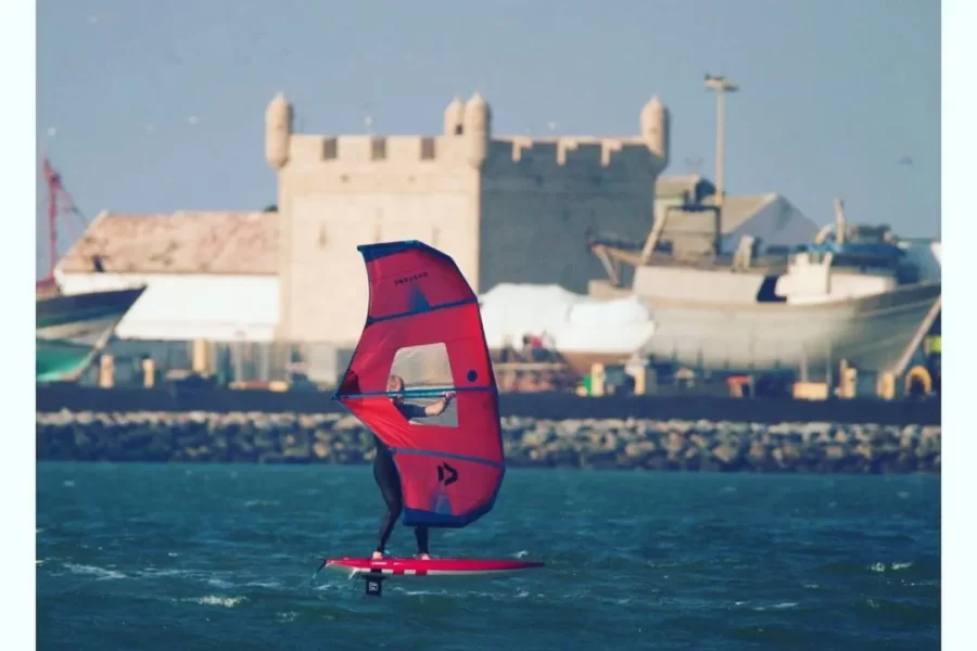 Single windsurfer on red sail in Essaouira bay with historic kasbah and fishing boats in background