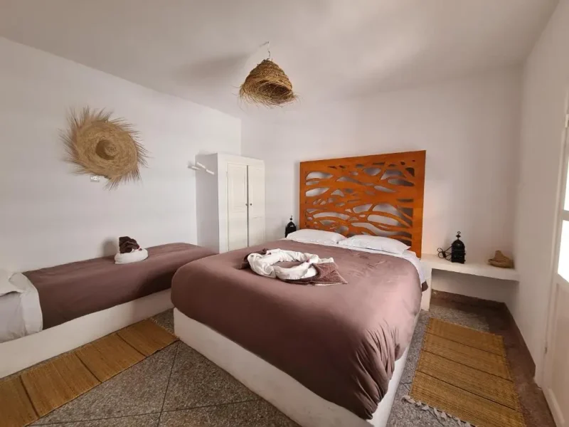 Chambre triple à Holy Beach House à Essaouira présente murs blancs, linge marron, tête de lit géométrique orange et décor tissé.