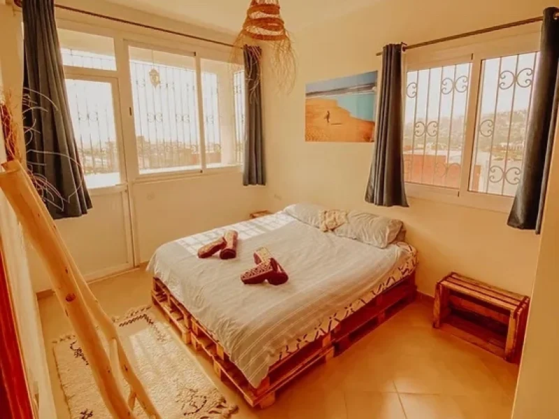 Double bedroom at Ohana Surf Morocco in Taghazout with white bedding, red wooden frame, and dual balcony access with ocean views.