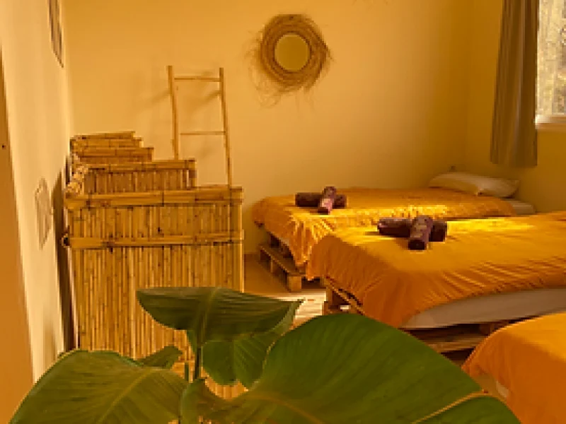 Yellow dormitory room with four beds covered in orange linens, woven wall decorations, and wooden storage.