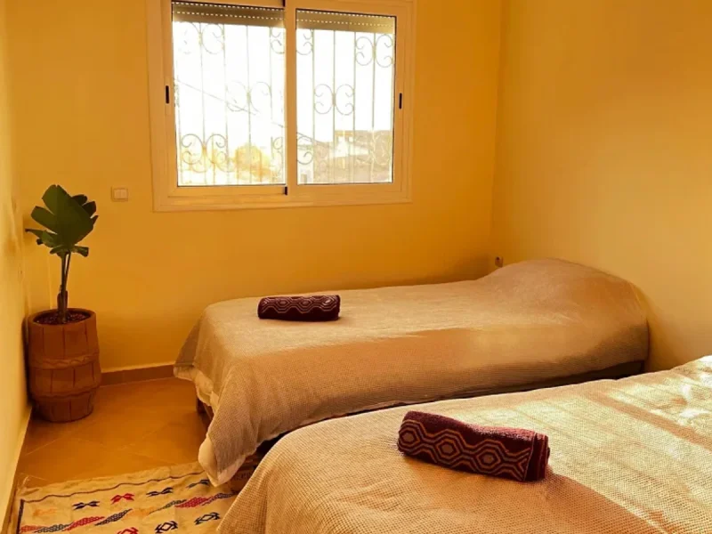 A bright yellow bedroom with two single beds, decorative maroon pillows, and tall windows with wrought iron grilles at Ohana Surf Morocco.