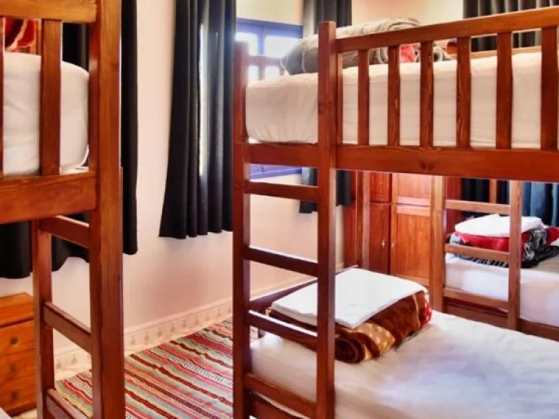 Six-bed shared dormitory room with wooden bunk beds, white mattresses, black curtain dividers, and striped rugs in Essaouira.