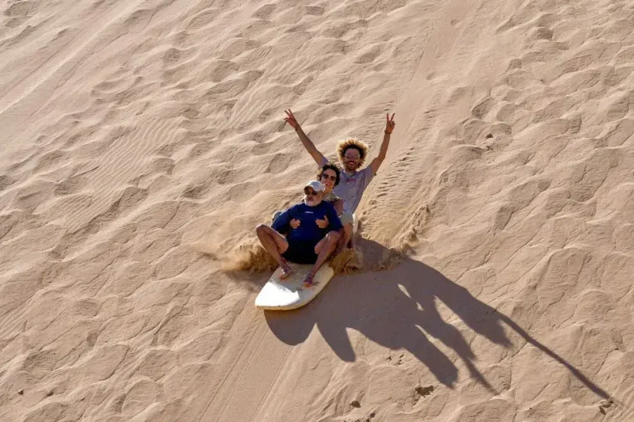 Three guests sandboard down a large sand dune in Taghazout while spectators watch from the crest above.