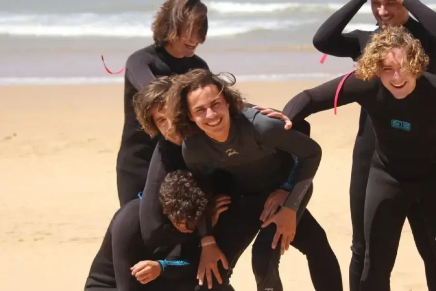 Cinq élèves de surf en combinaisons noires posant ensemble sur la plage de Sidi Kaouki à Essaouira avec vagues océaniques derrière eux.