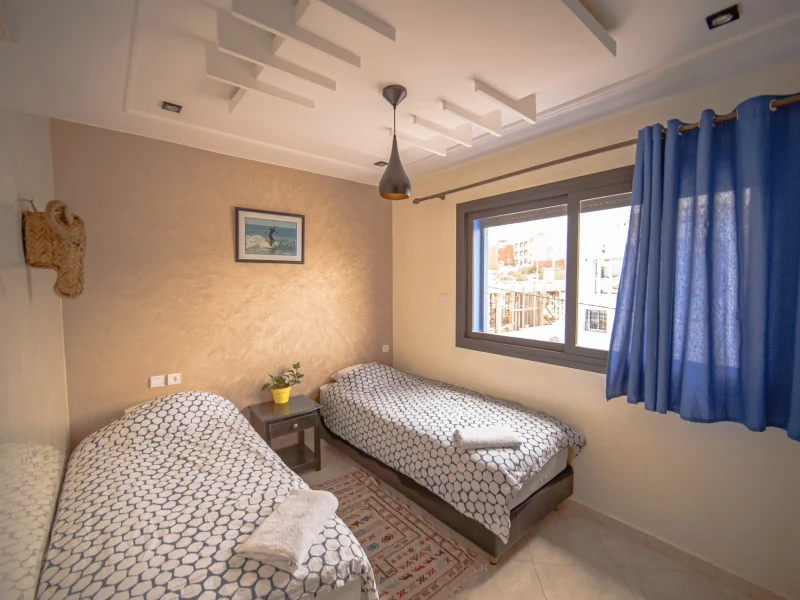 Private room with two single beds, geometric patterned bedding, and sea view from window in Imsouane.