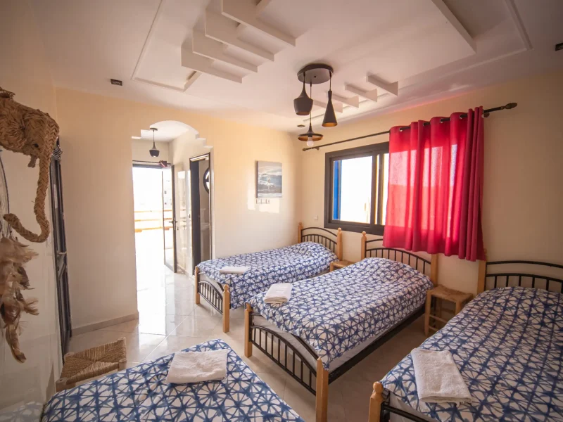 A bright shared bedroom in Imsouane featuring three single beds with patterned blue bedding, red curtains, and sliding door access to a balcony with ocean views.
