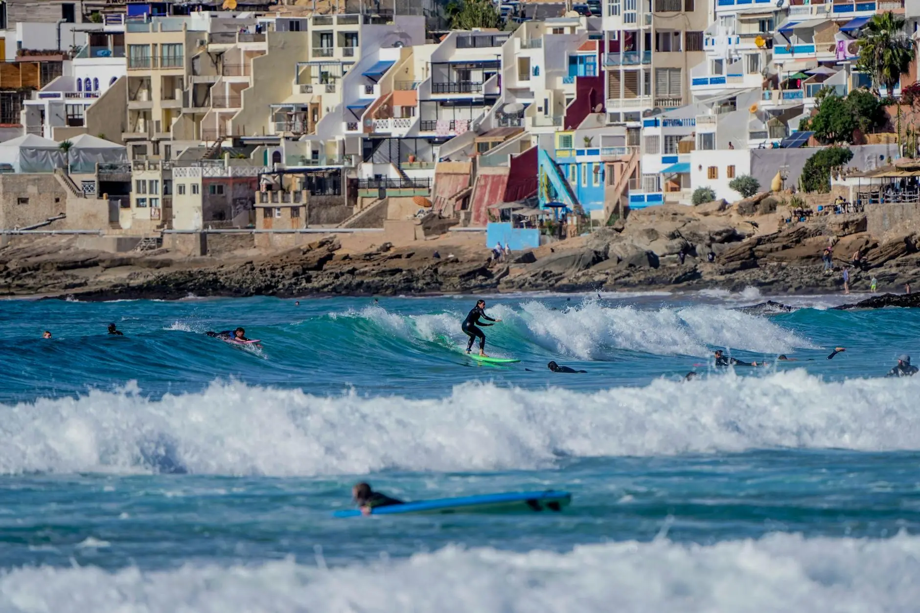 Mehrere Surfer im Wasser an einem marokkanischen Beachbreak mit weißen Häusern auf der Landzunge.