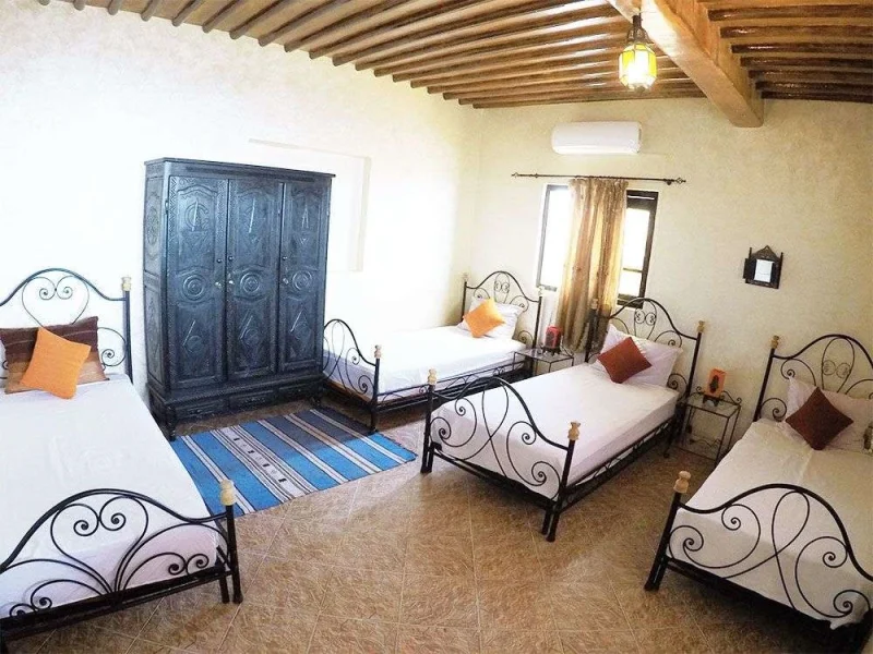 A shared dormitory room at Original Surf Morocco in Taghazout features multiple iron-framed beds with white linens and decorative cushions, a blue painted wardrobe, wooden ceiling beams, and woven striped rugs.