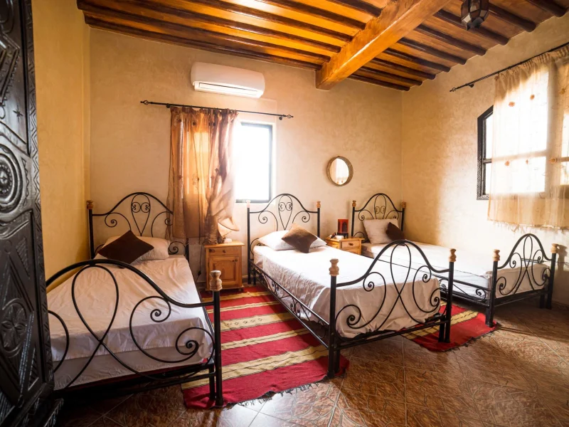 Triple room with three wrought iron beds, exposed wooden beams, and terracotta walls at Original Surf Morocco in Taghazout.
