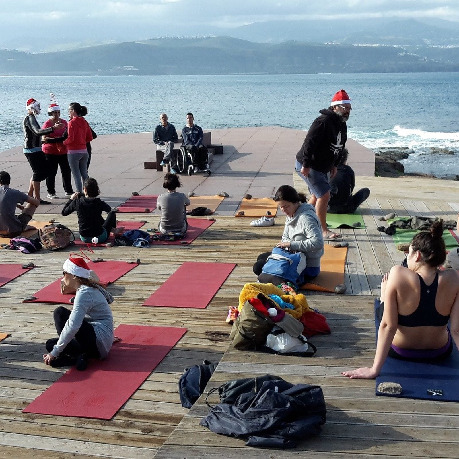 Gruppen-Yoga-Kurs auf einer Holzterrasse mit Blick auf den Ozean, mit Teilnehmern auf Yogamatten und sichtbaren Bergen über dem Wasser.