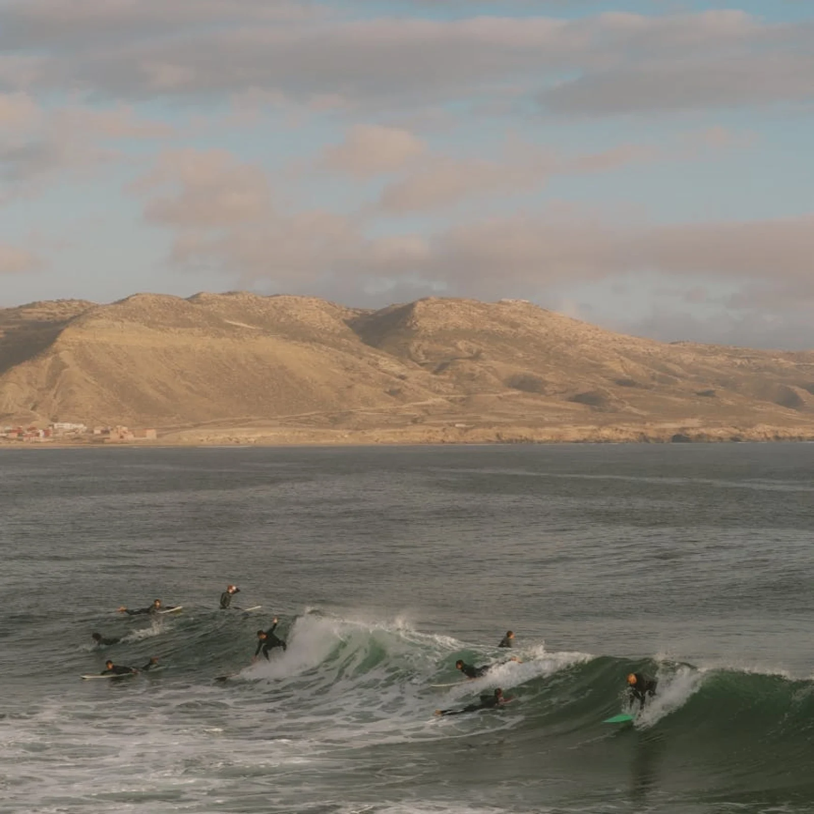Mehrere Surfer paddeln und reiten Wellen in der Bucht von Imsouane mit ariden Bergen im Hintergrund.