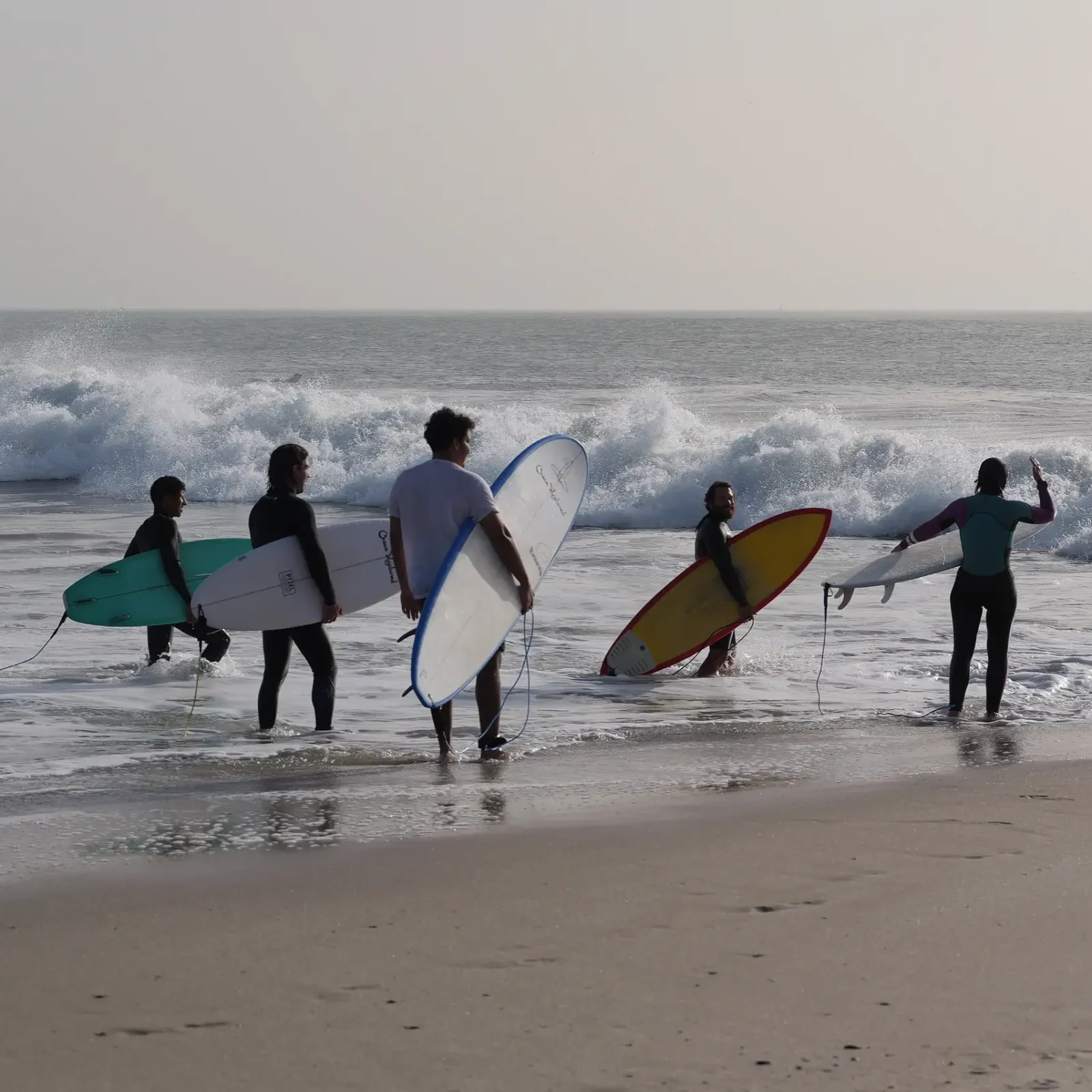 Group of five surfers in wetsuits carrying surfboards prepare to enter breaking waves on a Moroccan beach.