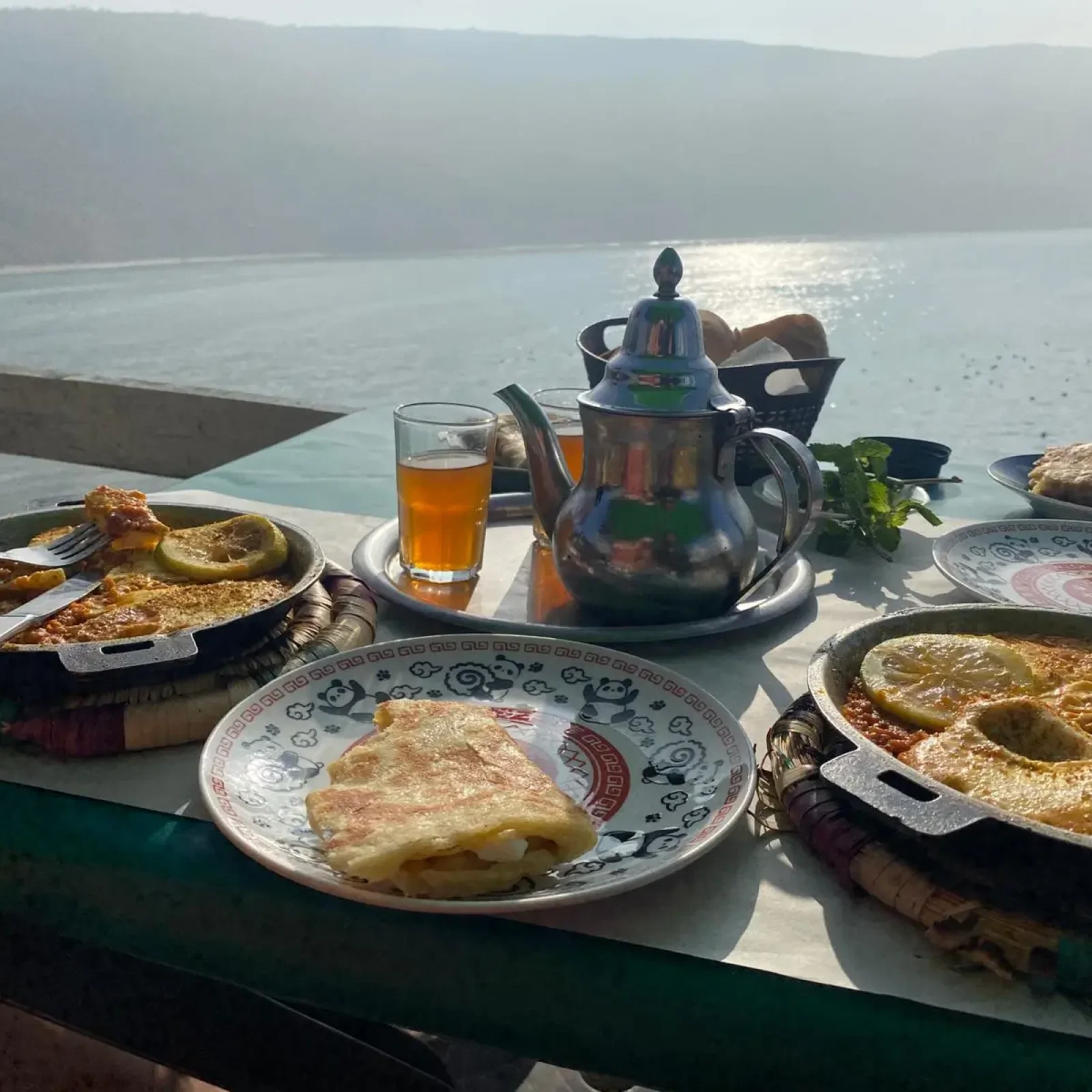 Uferpromenaden-Speisesetup in Imsouane mit Crêpes, dekorativen Tellern, traditioneller Metall-Teekanne und Teeglas mit Blick auf ruhige Buchtgewässer und umliegende Berge.