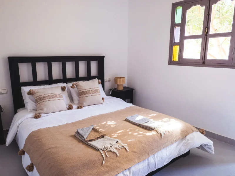 Queen Room bed with black headboard, beige and cream bedding with decorative cushions, at Surf Les Vagues in Sidi Kaouki, Essaouira.