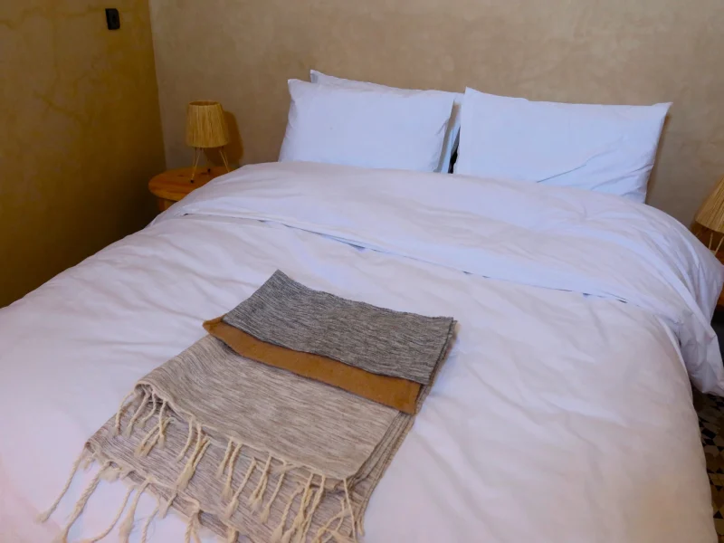 Double room with white linens and folded woven blankets on the bed, wooden side table with lamp visible.