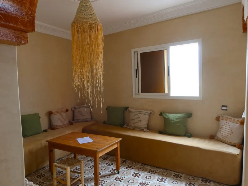 Interior of a room featuring a wooden table, built-in bench seating with patterned cushions, and a woven pendant light fixture.