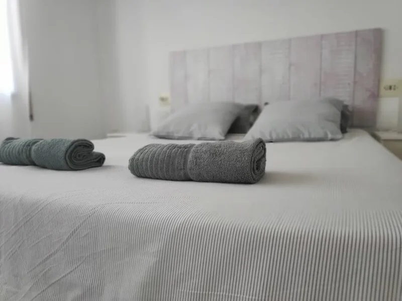 Gerollte graue Handtücher auf einem weißen Doppelbett mit Kissen und sichtbarem Kopfteil.