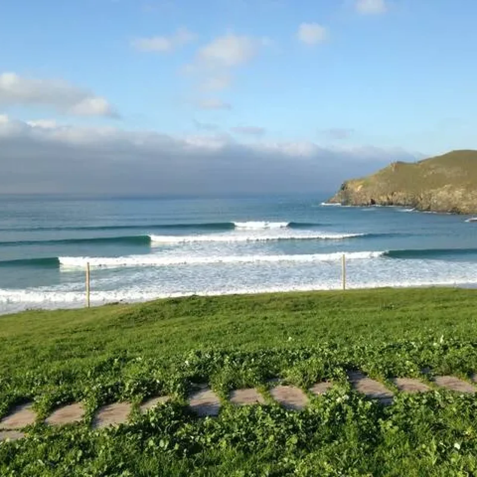 Pantin-Strand mit rollenden Wellen und Grasrasen, hinterlegt von felsigen Vorgebirgen unter teilweise bewölktem Himmel.