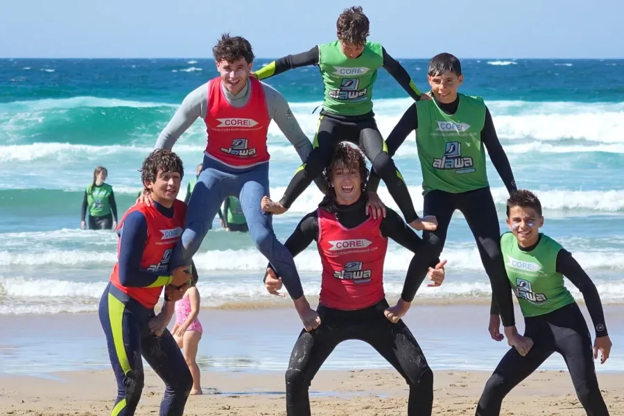 Gruppe von Kindern und Teenagern in roten und grünen Core- und Sawa-Neoprenanzügen posieren gemeinsam auf dem Sand am Pantin-Strand mit Wellen im Hintergrund.