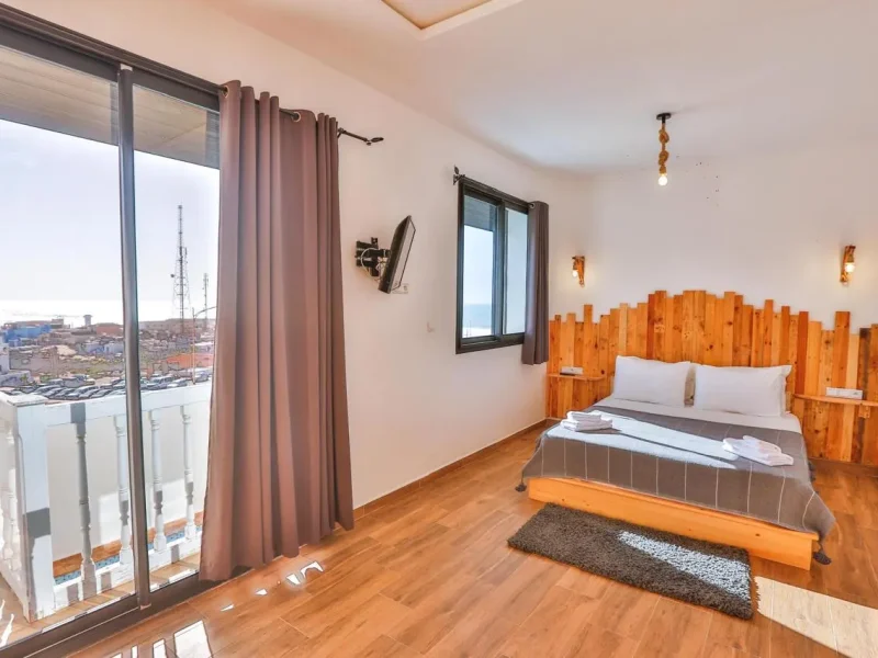Familienzimmer mit Holzkopfteil, Stadtblicken durch Balkontüren und Bettsitz im Moon Surf House in Imsouane.
