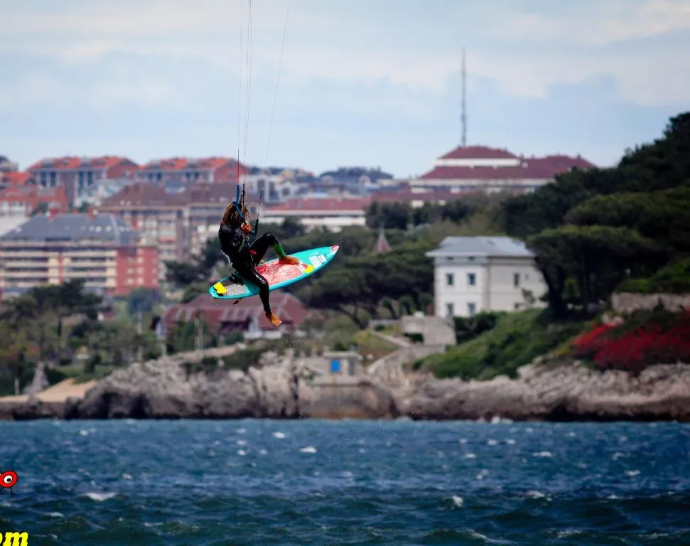 A kitesurfer executes an aerial trick with a colorful board above the rocky coastline near Cantabria, Spain, with residential buildings visible on the hillside.