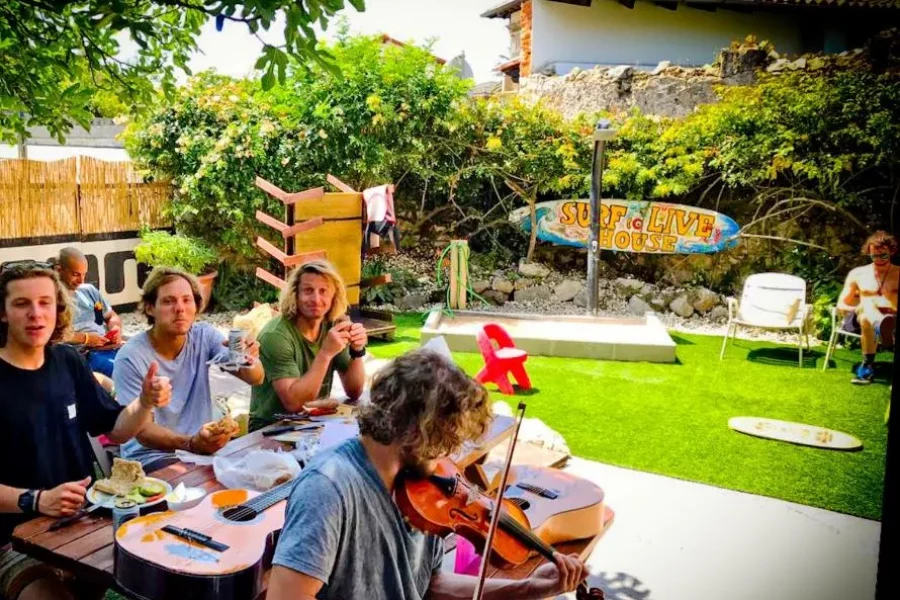 Gruppo di ospiti a pranzo e socializza sulla terrazza del giardino del Surf To Live con segnaletica visibile e vegetazione lussureggiante circostante.