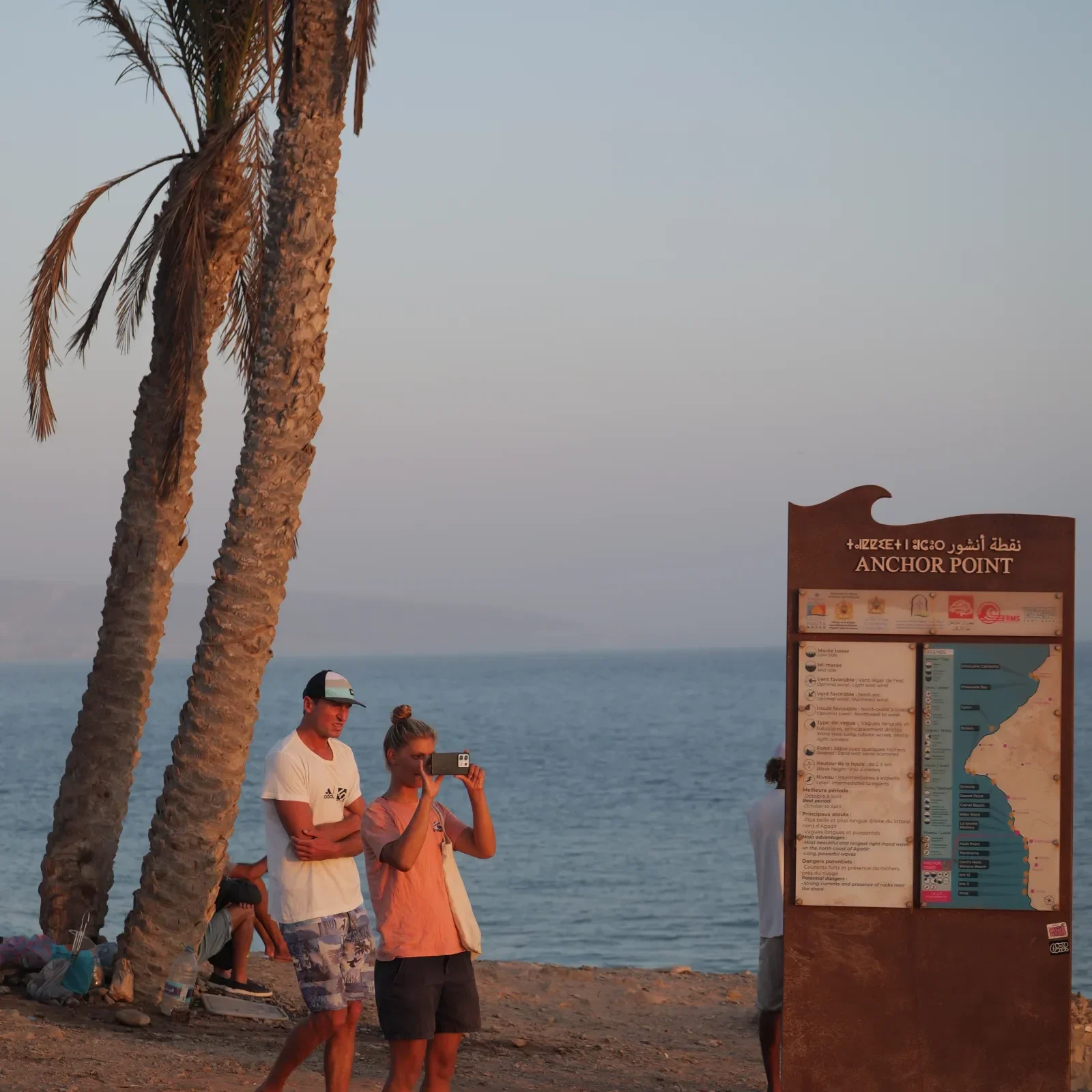 Besucher fotografieren den Ozean vom Strand Anchor Point, einem Küsten-Wahrzeichen in Marokko mit Palmen und Beschilderung.